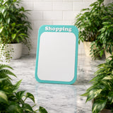 Teal-framed whiteboard with 'Shopping' text on a marble surface surrounded by plants.
