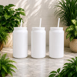 Three white mason jar-style tumblers with straws on a marble surface surrounded by plants.