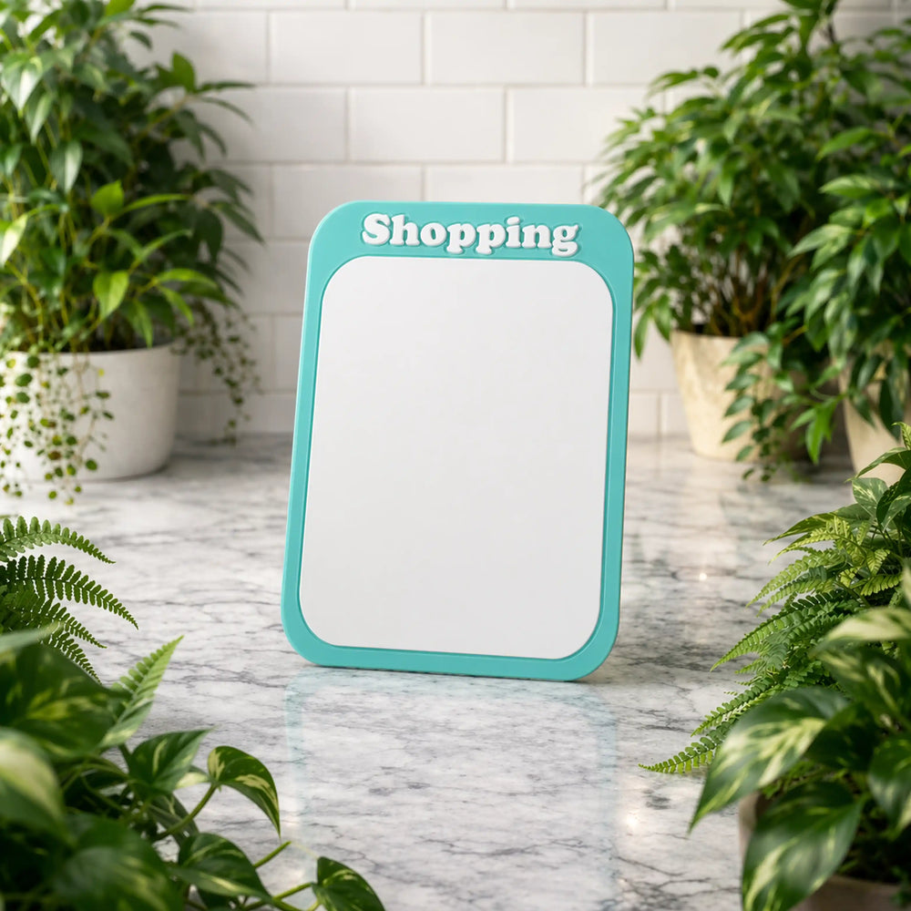 Teal-framed whiteboard with 'Shopping' text on a marble surface surrounded by plants.