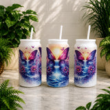 Three can-shaped tumblers with butterfly designs on a wooden surface with plants in the background.