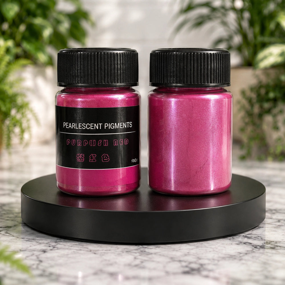 Two containers of purplish red pearlizing pigments on a black stand with a blurred green plant background.