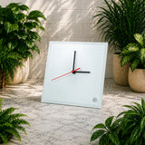 White square wall clock with black and red hands on a marble surface surrounded by potted plants.